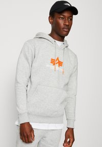 Felpa grigia con tasca frontale, con logo a stella arancione e testo "Alpha Industries". Indossata sopra una maglietta bianca e abbinata a dei pantaloni.