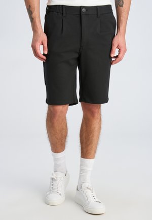 Modello maschile che indossa pantaloncini neri fino al ginocchio, calzini bianchi alti e sneakers bianche, in piedi con le mani rilassate lungo i fianchi.