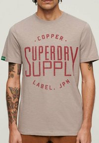Camiseta de algodón beige con un atrevido estampado gráfico rojo "SUPERDRY SUPPLY". Manga corta, cuello redondo, textura suave. Acentos mínimos.