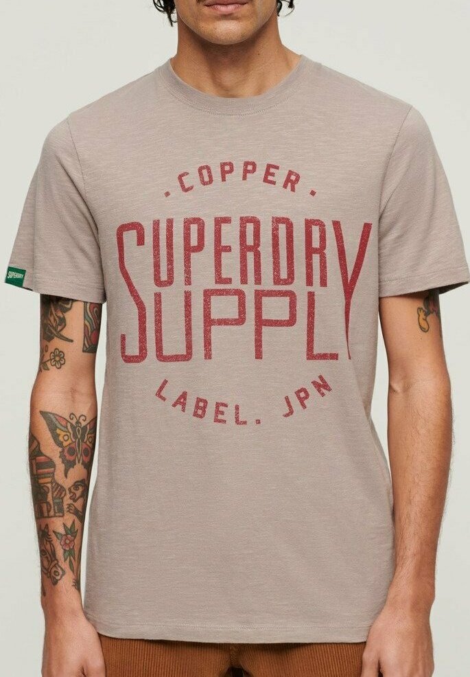 Camiseta de algodón beige con un atrevido estampado gráfico rojo "SUPERDRY SUPPLY". Manga corta, cuello redondo, textura suave. Acentos mínimos.