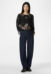 Žena v černém květinovém krajkovém topu s dlouhým rukávem, tmavých širokých džínách a černých teniskách, stojící před jednobarevným bílým pozadím.