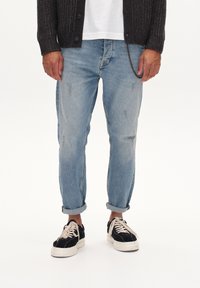 Lichtblauwe denim jeans met zichtbare slijtage, opgerolde mouwen, gecombineerd met zwarte suède schoenen met witte zolen en veters.