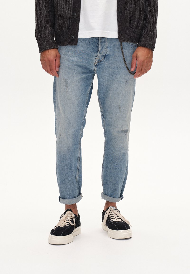 Lichtblauwe denim jeans met zichtbare slijtage, opgerolde mouwen, gecombineerd met zwarte suède schoenen met witte zolen en veters.