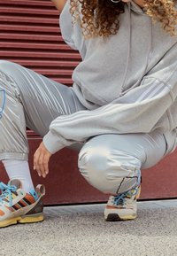 Sweat à capuche gris avec détail de cordon de serrage, joggers métalliques argentés avec rayures, et baskets multicolores présentant des couches et des accents texturés.