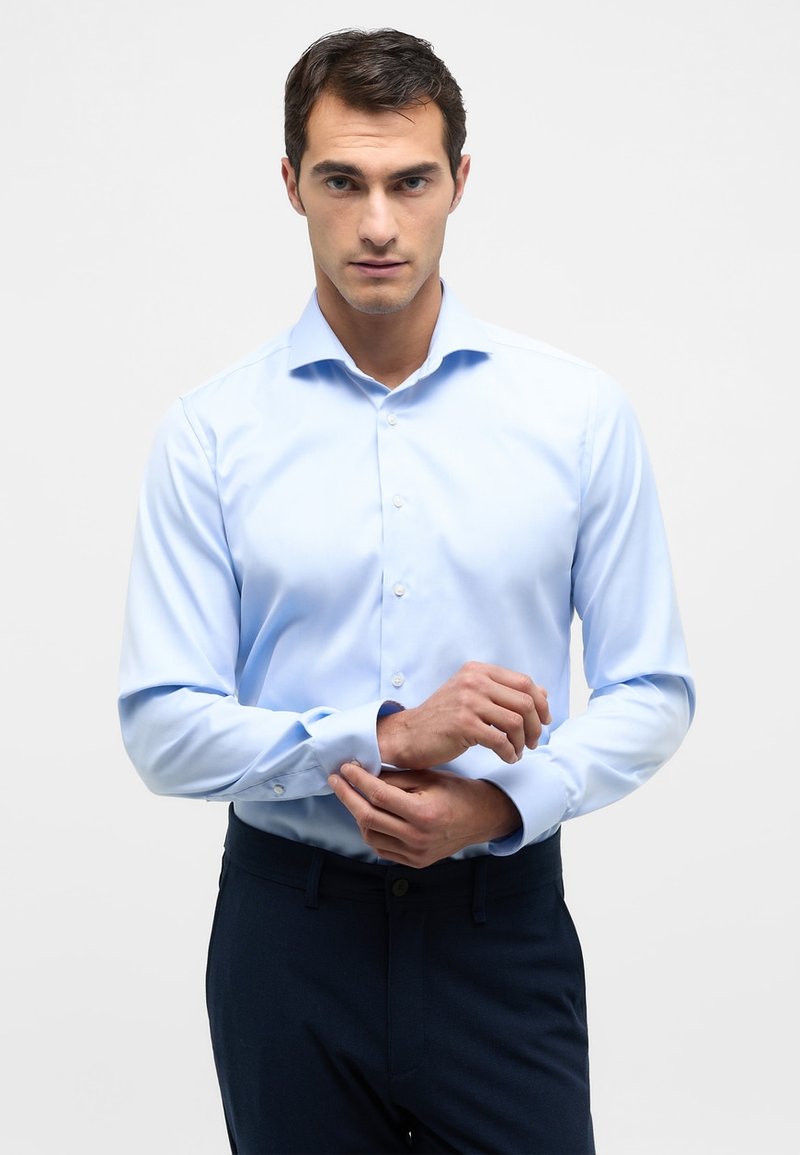 Lichtblauwe button-up shirt met lange mouwen, een kraag en een gladde textuur. Model past de manchet aan. Gedragen met donkere broeken.