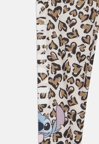 Beige háttérrel ellátott leggings, barna és fekete szívecskékkel és leopárd foltokkal. Kék színű Stitch grafika látható, pink nyelvvel.