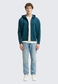 Joven de pie vistiendo una sudadera con cremallera color teal, camiseta blanca, jeans azul claro y zapatillas blancas frente a un fondo claro.