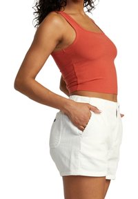 Kobieta ubrana w pomarańczowy, prążkowany top na ramiączkach i białe szorty z korduroy, z rękami w kieszeniach, pokazana z boku.