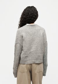 Άτομο με μακριά σγουρά μαλλιά γυρισμένο πλάτη, φοράει γκρι πλεκτό πουλόβερ με μανίκια που κλείνουν με κουμπιά και μπεζ παντελόνι με πίσω τσέπες.