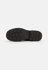 Sole di una scarpa da ginnastica nera con scanalature profonde, punta arrotondata e superficie testurizzata. Presenta un logo e due frecce nel design della suola.