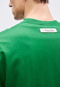 T-shirt verde de algodão com decote redondo e um rótulo da marca em branco na parte de trás do decote, exibindo uma textura suave e um design simples.