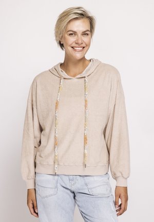 Lächelnde Frau mit kurzem blondem Haar trägt einen beigen Kapuzenpullover mit mehrfarbigen Kordeln und hellblauen Jeans, steht vor weißem Hintergrund.