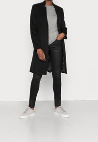 Schwarzer langer Mantel mit geradem Design, kombiniert mit einem grauen strukturierten Pullover und schwarzen Lederhosen. Weiße Sneaker mit strukturiertem Obermaterial.