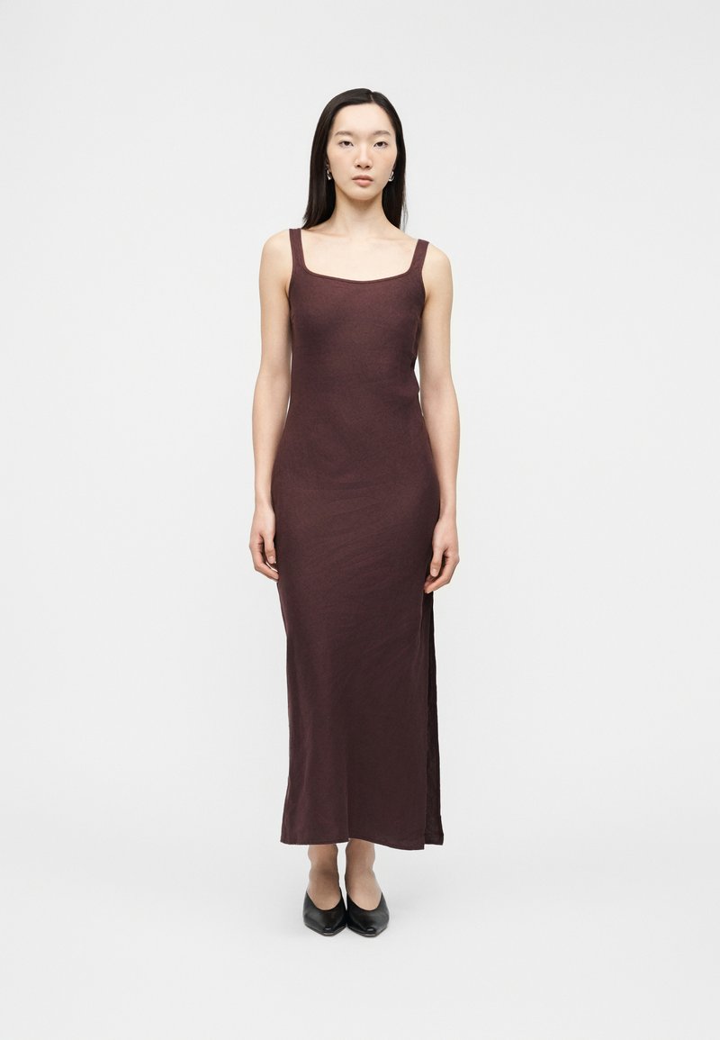 Γυναίκα με ίσια μαύρα μαλλιά που φοράει αμάνικο, μακρύ φόρεμα σκούρου καφέ χρώματος με τετράγωνη λαιμόκοψη και μαύρες μπαλαρίνες, στέκεται μπροστά σε ένα απλό λευκό φόντο.