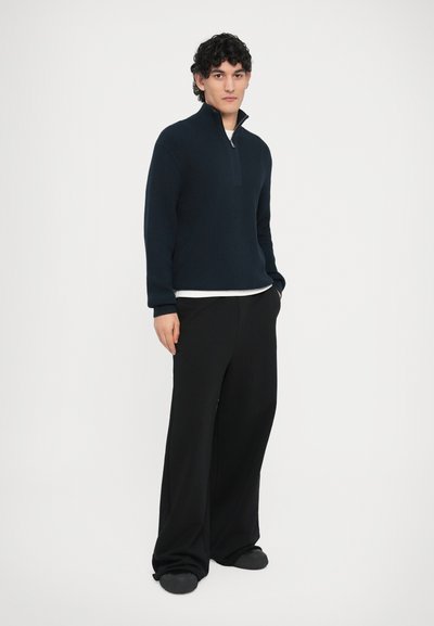 Pull-over en maille bleu marine avec col montant et demi-fermeture éclair, associé à un pantalon noir ample et des chaussures foncées. Design simple et fonctionnel.