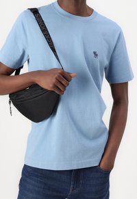 Hombre vistiendo una camiseta de color azul claro con un pequeño logo bordado, jeans oscuros y sosteniendo un bolso cruzado negro con una correa de marca.