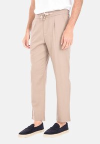 Pantaloni tapered beige, leggeri, con elastico in vita, cordino e due tasche laterali; texture liscia e design con pieghe frontali.