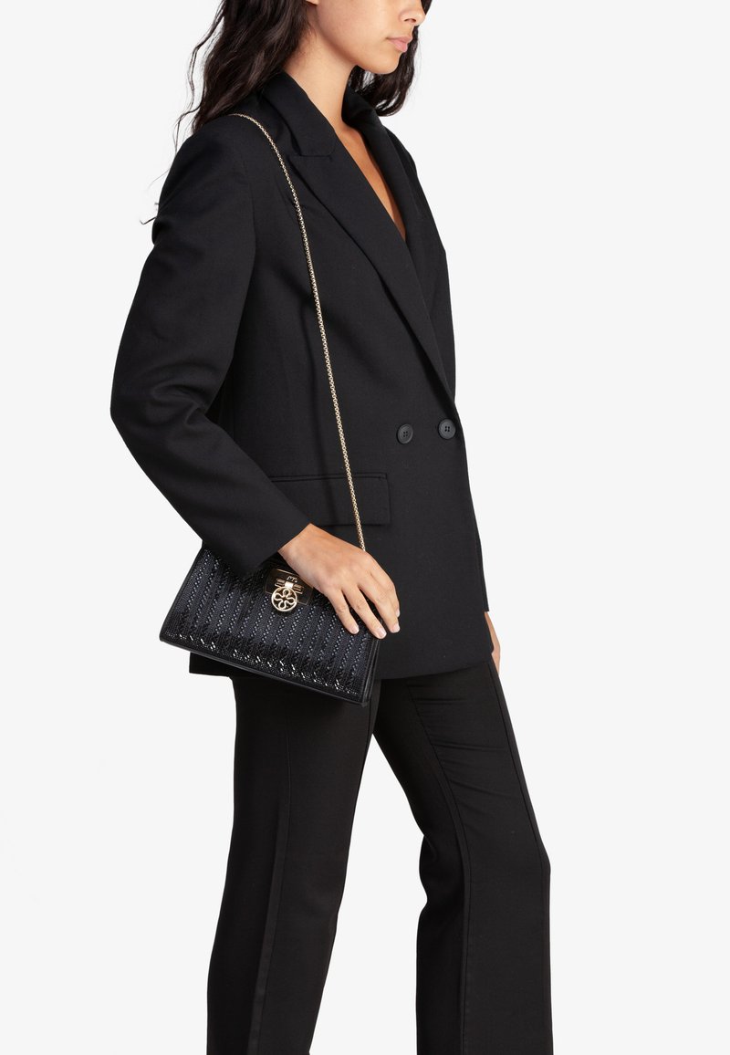 Borsa a tracolla nera trapuntata con hardware dorato, tracolla a catena e superficie texturizzata. Abbinata a un blazer nero su misura e pantaloni.