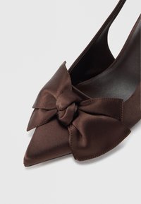 Zapato de tacón alto de satén marrón con tira trasera y punta puntiaguda, adornado con un gran lazo decorativo en la parte delantera. Textura suave con acabado brillante.