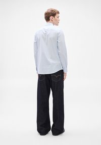 Camisa de rayas azul claro de manga larga con cuello clásico, combinada con vaqueros oscuros de pierna ancha. Vista desde atrás, mostrando la longitud de la camisa.