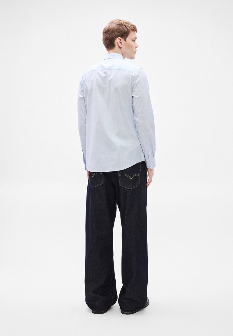 Hellblau gestreiftes Langarmhemd mit klassischem Kragen, getragen zu dunklen, weit geschnittenen Jeans. Ansicht von hinten, um die Länge des Hemdes zu zeigen.