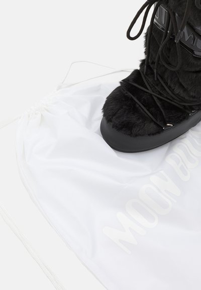 Botte d'hiver en fourrure noire avec une semelle noire brillante, associée à un sac à cordon blanc portant le texte "MOON BOOTS" dans un imprimé subtil.