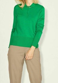 Person klädd i en klargrön långärmad pikétröja och beiga byxor, stående mot en enkel ljus bakgrund.