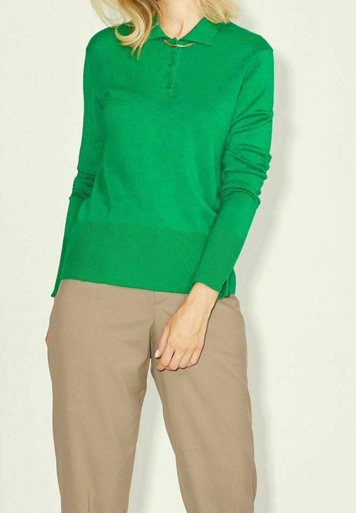 Person klädd i en klargrön långärmad pikétröja och beiga byxor, stående mot en enkel ljus bakgrund.