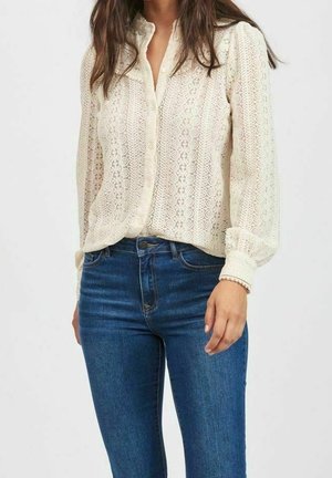 Femme portant un chemisier en dentelle crème à manches longues rentrée dans un jean en denim bleu, debout devant un fond clair uni.