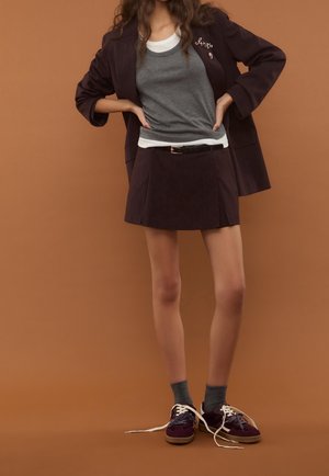 Débardeur gris superposé à une chemise blanche, blazer à fines rayures foncées, et mini-jupe plissée assortie. Porté avec des baskets violettes et des chaussettes grises.