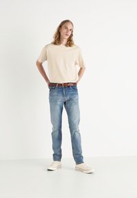 Helles beige Rundhals-T-Shirt, mittelblaue Jeans, brauer Gürtel mit Muster und weiße Sneakers. Model steht mit den Händen in den Hüften vor einem weißen Hintergrund.