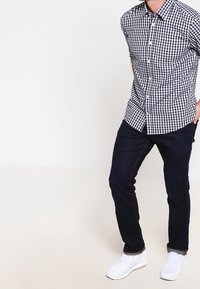 Uomo che indossa una camicia a scacchi bianco e nero, jeans blu scuro e sneaker bianche, in piedi con le mani dietro la schiena su uno sfondo bianco.