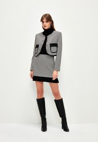 Mustat ja valkoiset houndstooth-kuvioiset takki, jossa on kaksi taskua, yhdistettynä mustaan poolopaitaan ja yhteensopivaan hameeseen. Polven ylle yltävät mustat nahkasaappaat.