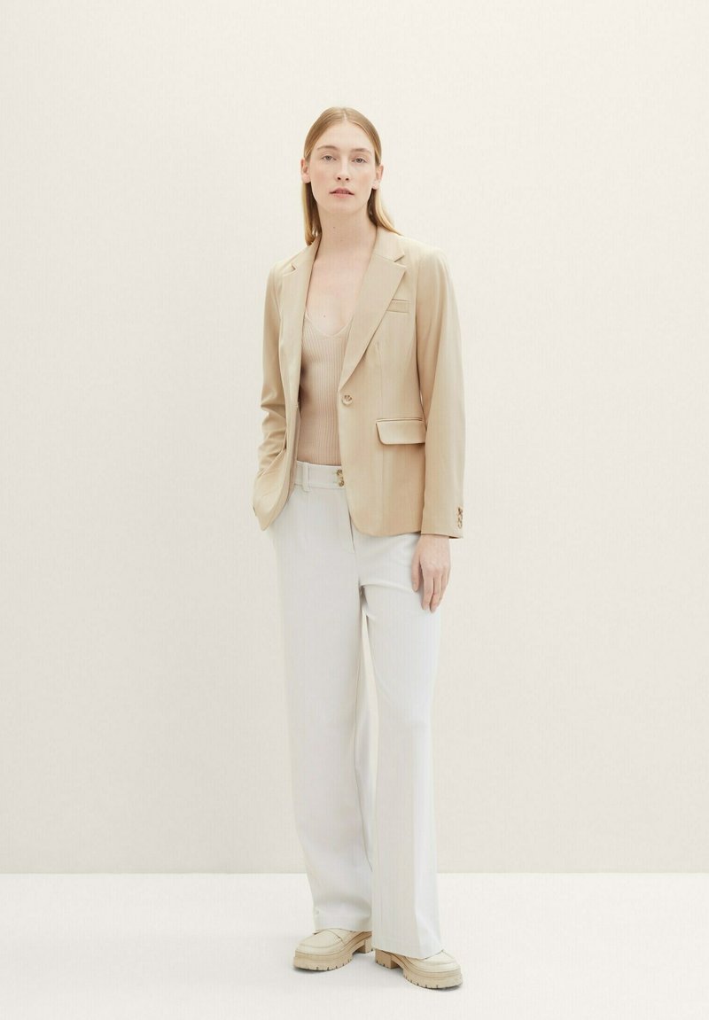 Blazer beige con un bottone, abbinato a un top senza maniche beige chiaro, pantaloni bianchi a gamba larga e scarpe beige con platform. Dettagli in tessuto testurizzato.