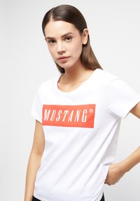 Camiseta de algodón blanca con un logotipo rectangular rojo prominente que dice "MUSTANG" en texto blanco. Con mangas cortas y un cuello redondo.