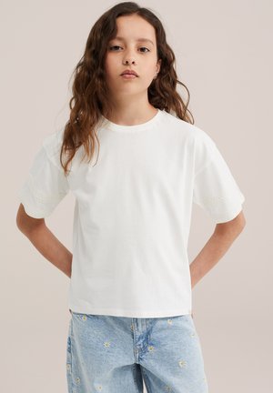 Pige med langt bølget hår iført en hvid t-shirt med blonder og lyseblå jeans pyntet med små hvide tusindfryd.