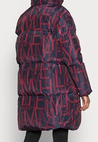 Chaqueta de plumas azul marino con un patrón de texto gráfico rojo, diseño acolchado, cuello alto y una silueta hasta la rodilla. Presenta puños elásticos y capucha.