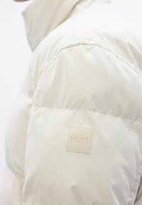 Gros plan d'une manche de doudoune beige clair avec une étiquette carrée cousue portant la mention « BOSS ».