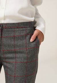 Pantaloni grigi a quadri con sottili motivi a scacchi neri e rossi, caratterizzati da una texture liscia e una tasca laterale. Abbinati a una camicia bianca.