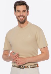 Beige tričko s krátkým rukávem a kulatým výstřihem, s jemným logem v texturované tkanině; modelka má na sobě vzorovaný opasek a světlé kalhoty.