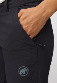 Svarte turbukser har en sidelomme, en elastisk snøring, og en lyseblå logo av et mammut på venstre lår.
