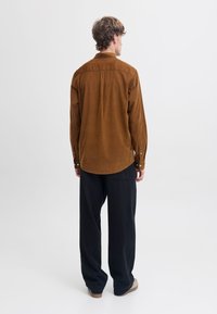 Brun manchester skjorta med rak skärning, knäppta manschetter och rundad fåll, kombinerad med lösa svarta byxor och beige skor.