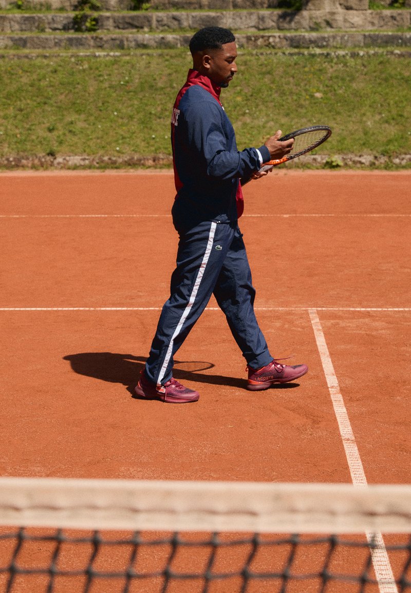Joueur de tennis masculin en survêtement bleu marine et chaussures rouges, tenant une raquette et marchant sur un court de tennis en terre battue près du filet.