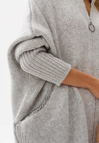 Szary sweter z dzianiny z zamkiem z przodu, w oversize'owym fasonie, z prążkowanymi rękawami i podszewką kieszeni w kratkę. Miękka textura i ciepły wygląd.