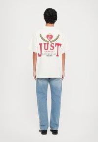 White oversized t-shirt with red graphics; features heart and laurel design, and text "JUST CAVALLI LOVERS CLUB Milano" on back.