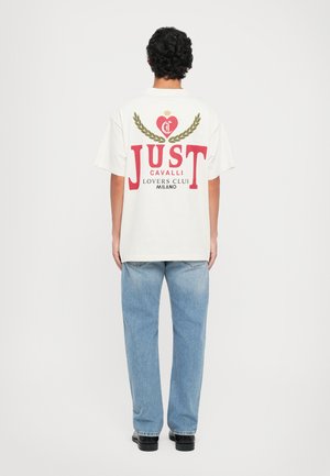 Camiseta blanca de gran tamaño con gráficos rojos; presenta un diseño de corazón y laurel, y el texto "JUST CAVALLI LOVERS CLUB Milano" en la parte trasera.