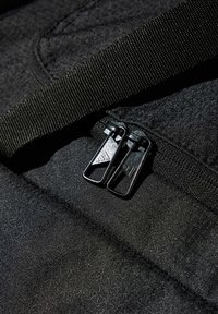 Mustalla kankaalla valmistettu reppu, jossa on tekstuuri, kestävät kudotut hihnat ja kaksinkertaiset mustat vetoketjut, joissa on kolmion muotoisia koristeita.