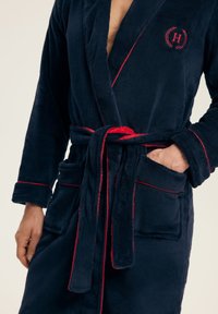 Granatowy szlafrok z pluszowego materiału z czerwonymi lamówkami, przewiązywany w talii i z dwoma przednimi kieszeniami; wyhaftowane logo na piersi.