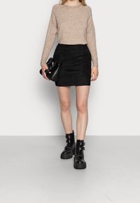 Pull en tricot beige, mini-jupe en suède noire et bottes noires volumineuses avec boucles. Portant un sac à main noir au design froncé.