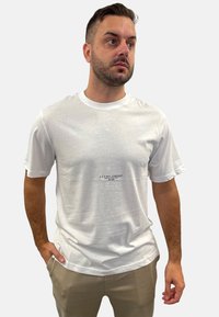 Weißes Baumwoll-T-Shirt mit kurzen Ärmeln und rundem Halsausschnitt. Verfügt über ein kleines schwarzes Logo auf der Brust. Glatte Textur.